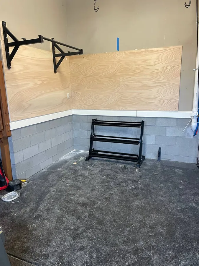 Home gym wall with a plywood backer and a pull-up bar mounted over block, rubber floor below